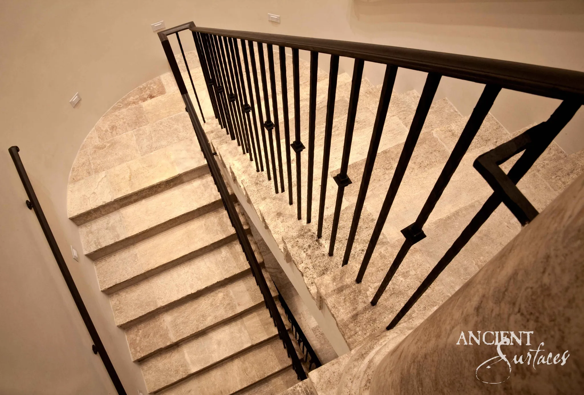 Reclaimed antique limestone stair from Ancient Surfaces photographed from the landing above, warm cream treads turning through a quarter landing beside an iron balustrade under a dark timber handrail
