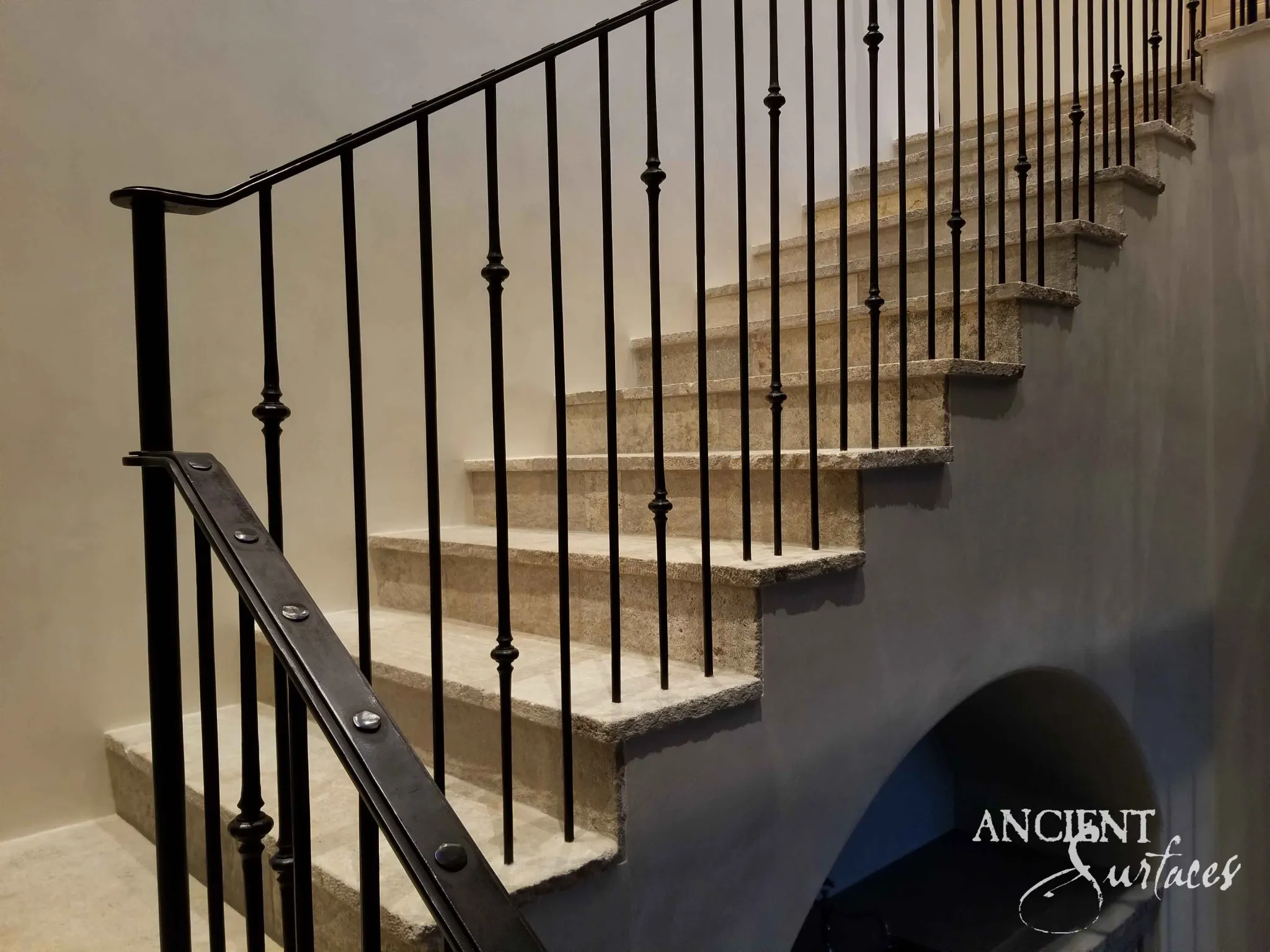 Side view of a reclaimed antique limestone stair from Ancient Surfaces with deep treads left in a coarse split face along their front edges, a turned iron balustrade, a riveted flat iron rail and an arched alcove beneath the flight