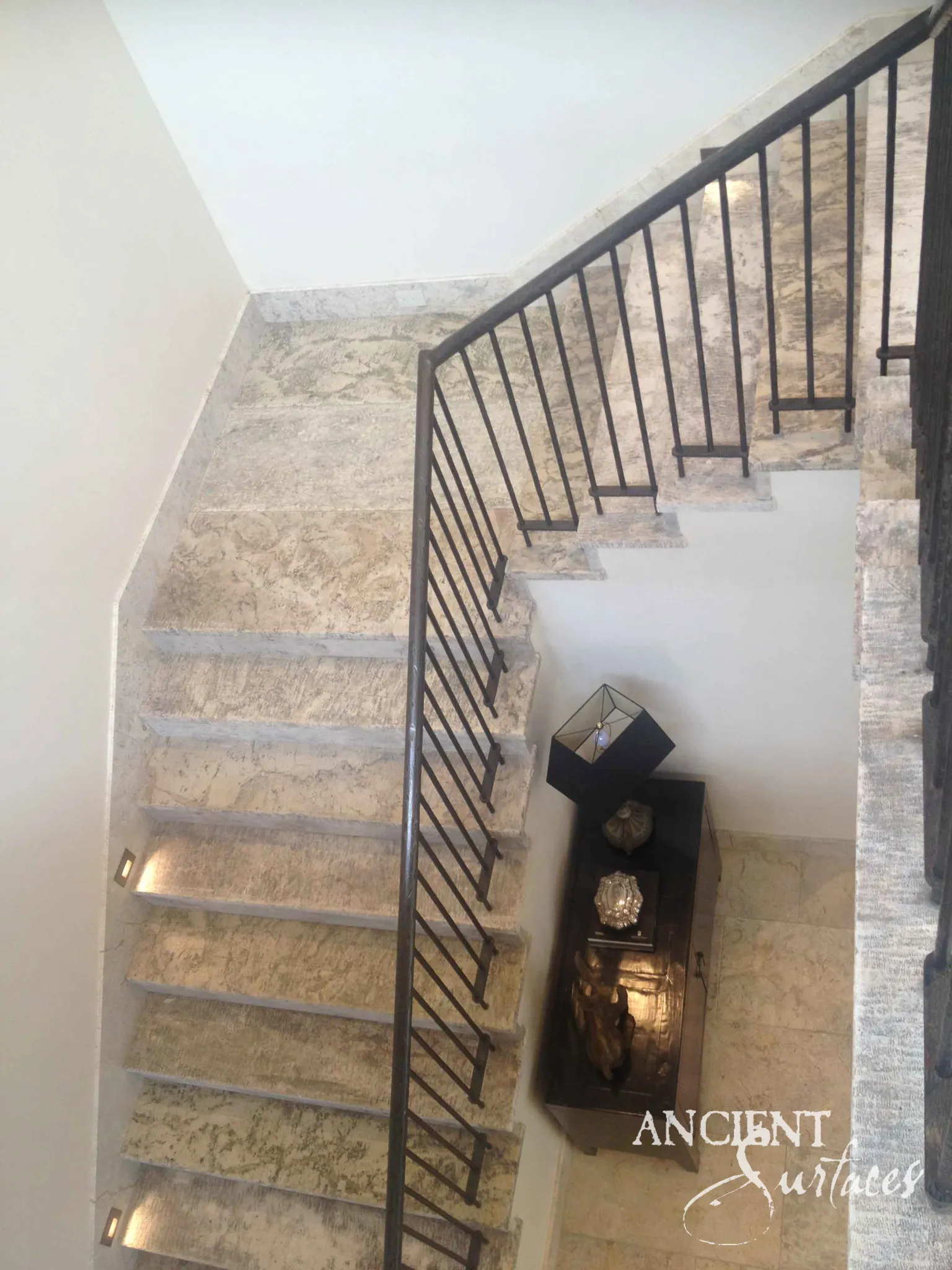 Looking down a reclaimed antique limestone stairwell from Ancient Surfaces, pale grey treads dense with fossil pockets and open pitting beside a minimal square bar iron railing