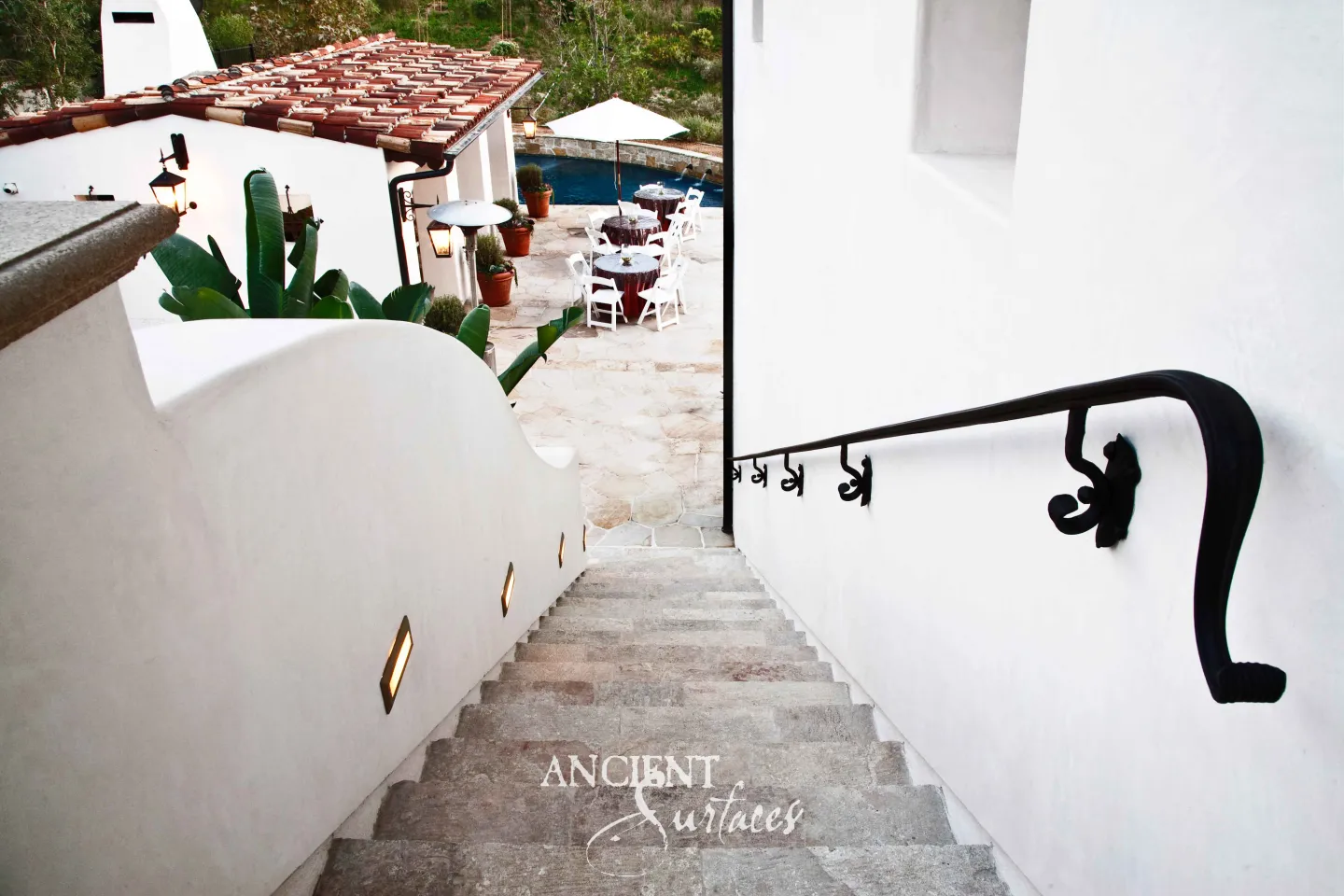 Reclaimed antique limestone garden steps from Ancient Surfaces descending between whitewashed walls toward a pool terrace, with scrolled wrought iron handrail brackets and recessed step lights