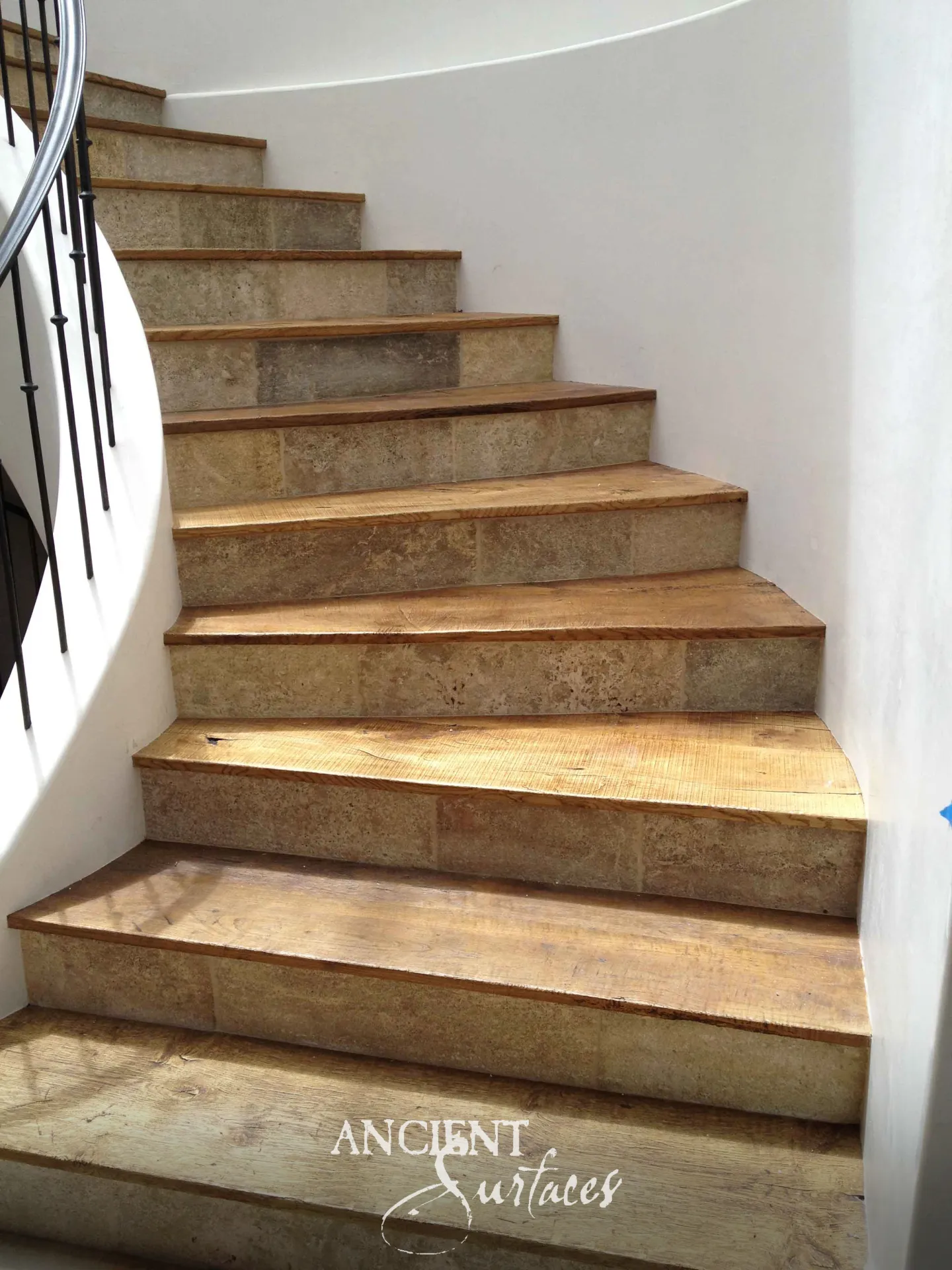 Winding stair from Ancient Surfaces with reclaimed antique limestone risers in mottled honey and olive stone beneath broad sawn timber treads, curving past a white plaster wall with an iron balustrade beyond