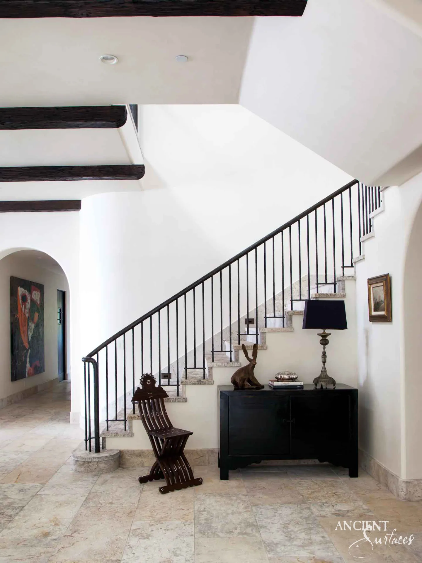 Reclaimed antique limestone stair from Ancient Surfaces climbing a stucco stringer in a double height villa hall under dark timber ceiling beams, with rough ended cream treads, a slim black iron balustrade and a rounded bottom step