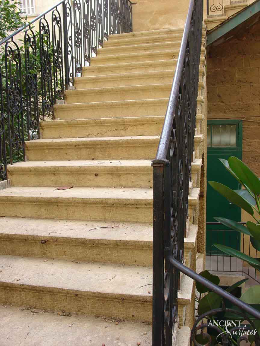 Reclaimed antique limestone stair from Ancient Surfaces in an old town courtyard, sandy gold treads dished hollow by use, ornate scrolled wrought iron balustrades on both sides and a green painted door under ochre plaster