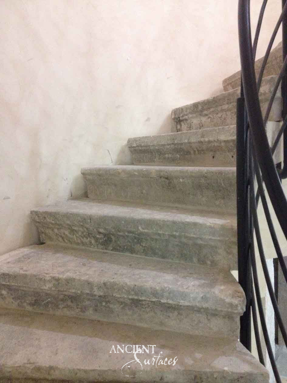 Reclaimed antique limestone stair from Ancient Surfaces against a limewashed wall, thick treads whose nosings have been rounded soft by centuries of footfall, the stone cast green grey