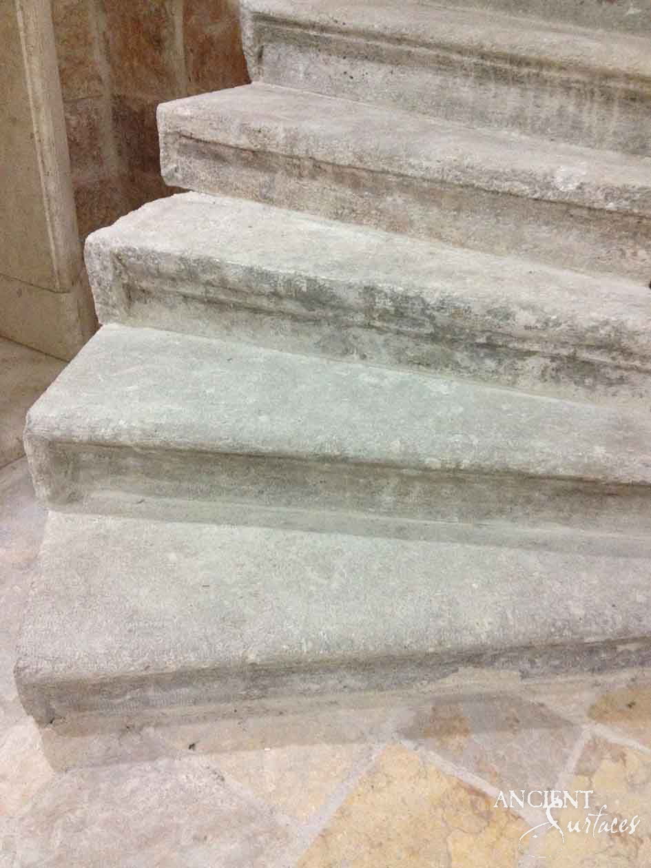 Close view of a reclaimed antique limestone stair from Ancient Surfaces turning past a plain stone jamb, full slab treads with square blunt nosings and a green grey bloom, landing on irregular honey toned flagstones
