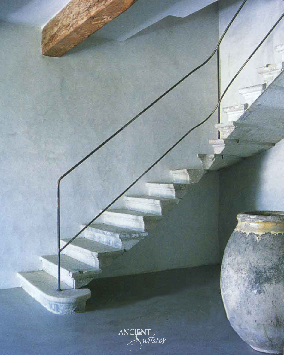 Reclaimed antique limestone stair from Ancient Surfaces with open string treads and moulded front edges floating against a blue grey plaster wall, a curved bottom step and a slim iron handrail beside a large glazed olive jar