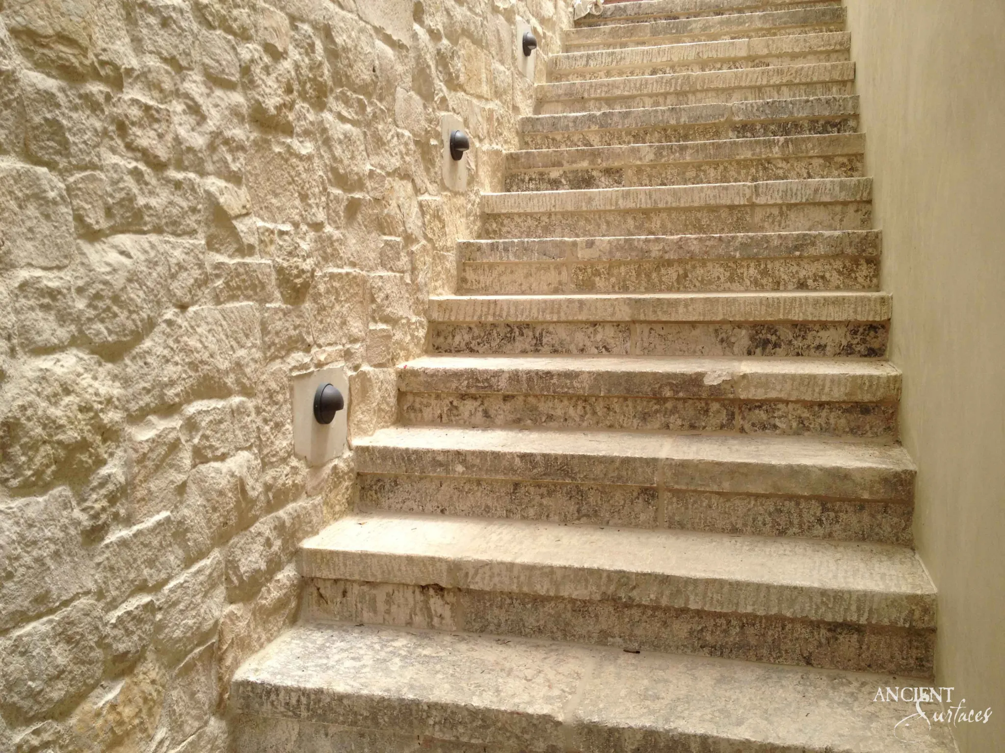 Reclaimed antique limestone stair flight from Ancient Surfaces climbing beside a rough coursed rubble limestone wall, the treads heavily chiselled with the tool marks still legible