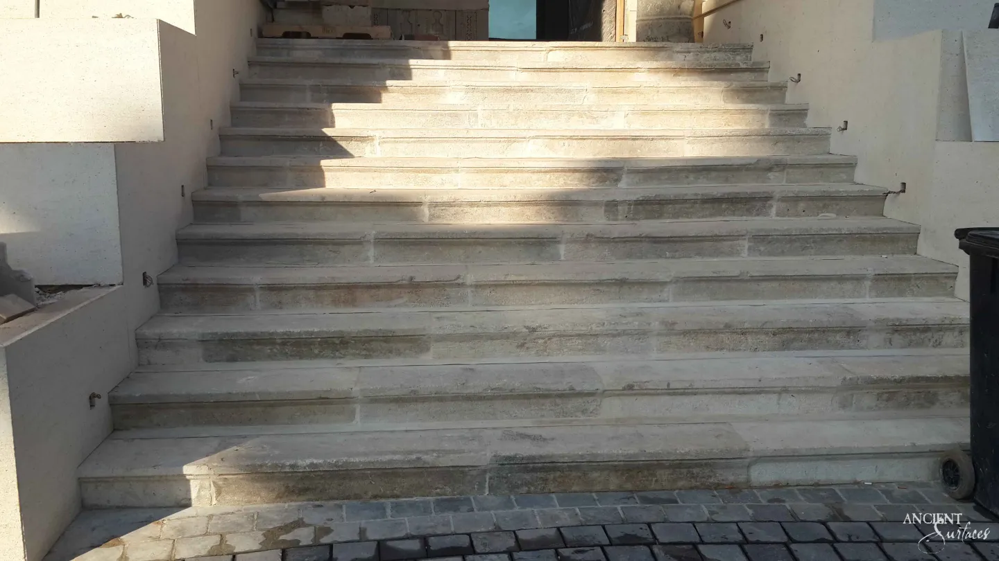 Broad shallow flight of reclaimed antique limestone entrance steps from Ancient Surfaces in cool pale cream striated stone, photographed in low sun during installation above freshly laid cobble paving