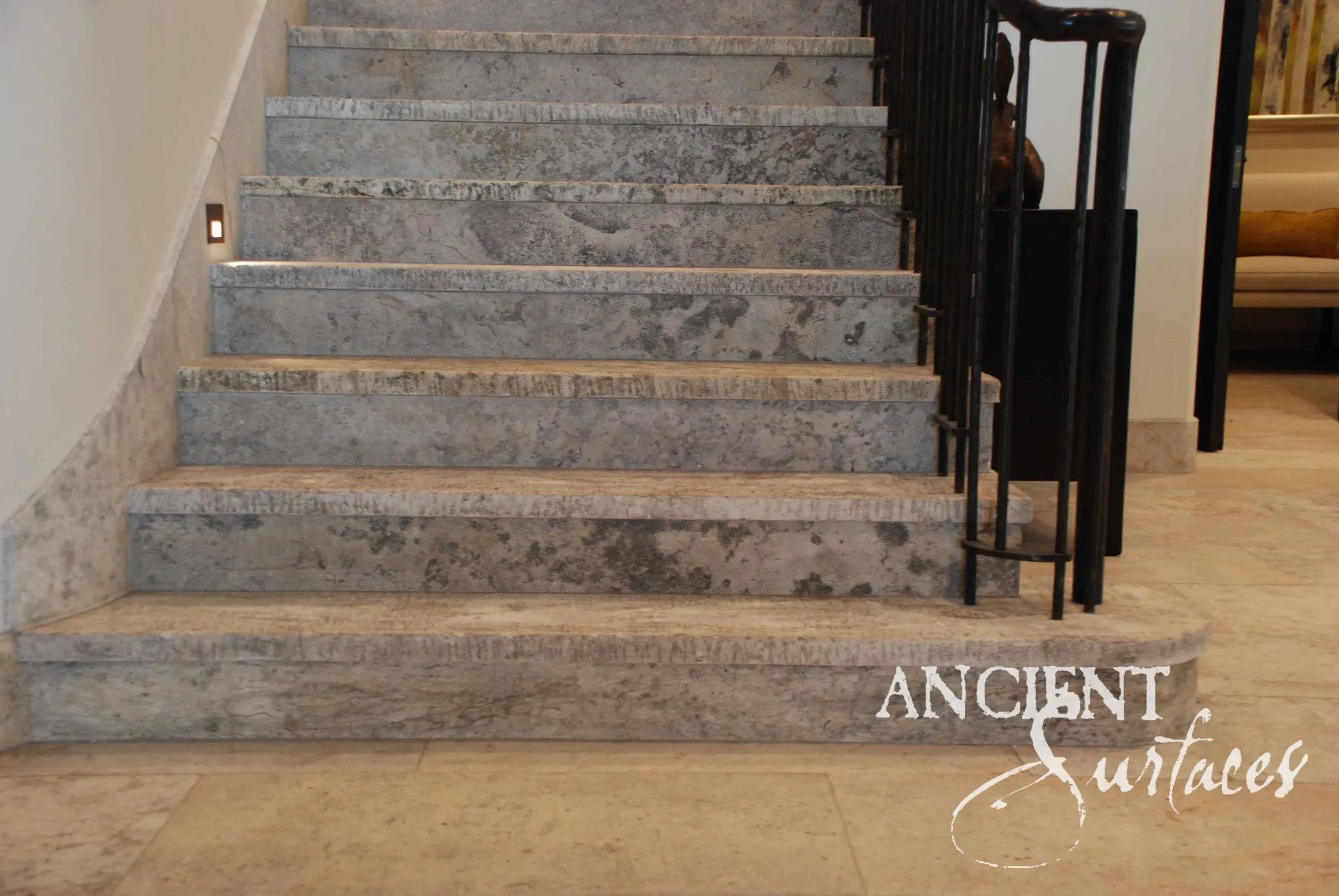 Foot of a reclaimed antique limestone stair from Ancient Surfaces in heavily veined silver grey stone, with a broad rounded starting step, a sculptural black iron newel and warm travertine paving alongside