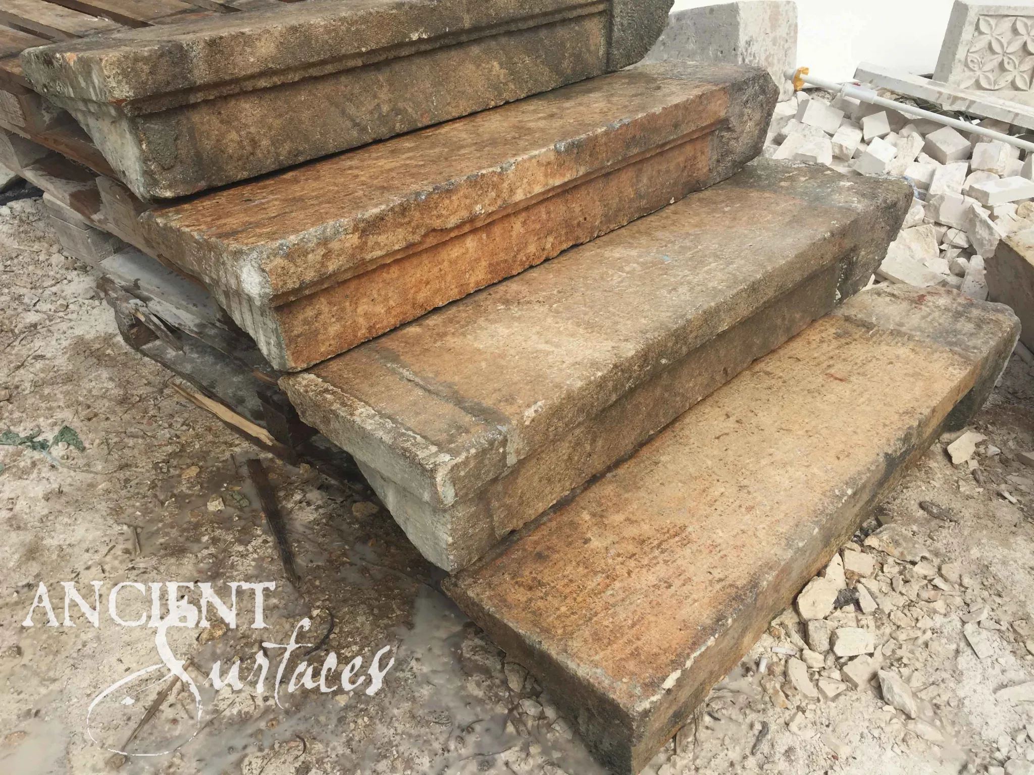 Reclaimed antique limestone steps from Ancient Surfaces stacked on a timber pallet in the stoneyard, the stone iron stained rust and ochre, front edges worked with a slim moulded drip, rubble and a carved rosette panel behind